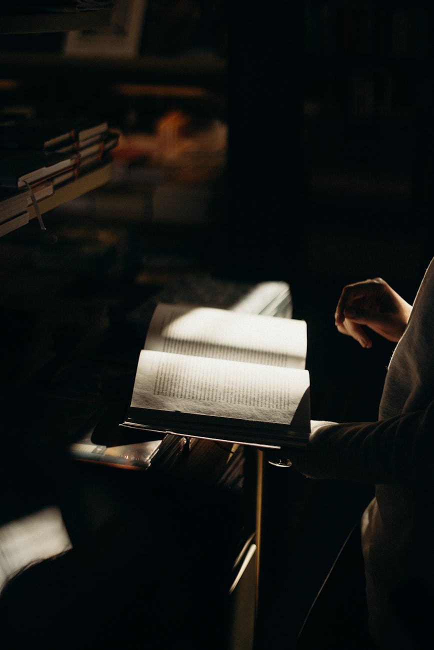 A dimly lit scene of a person reading an open book in a cozy, atmospheric space.