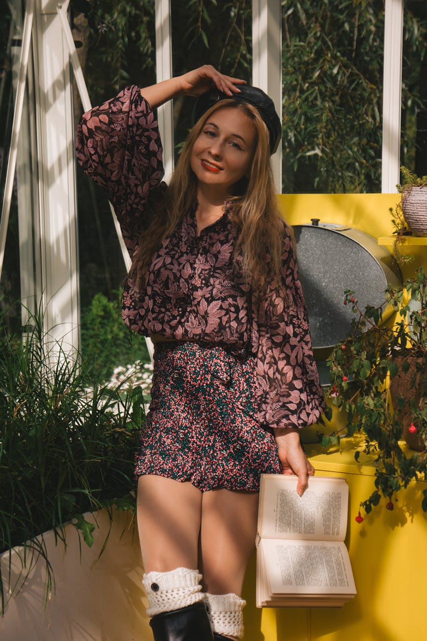 Woman in floral attire posing indoors holding a book with plants and a yellow backdrop.