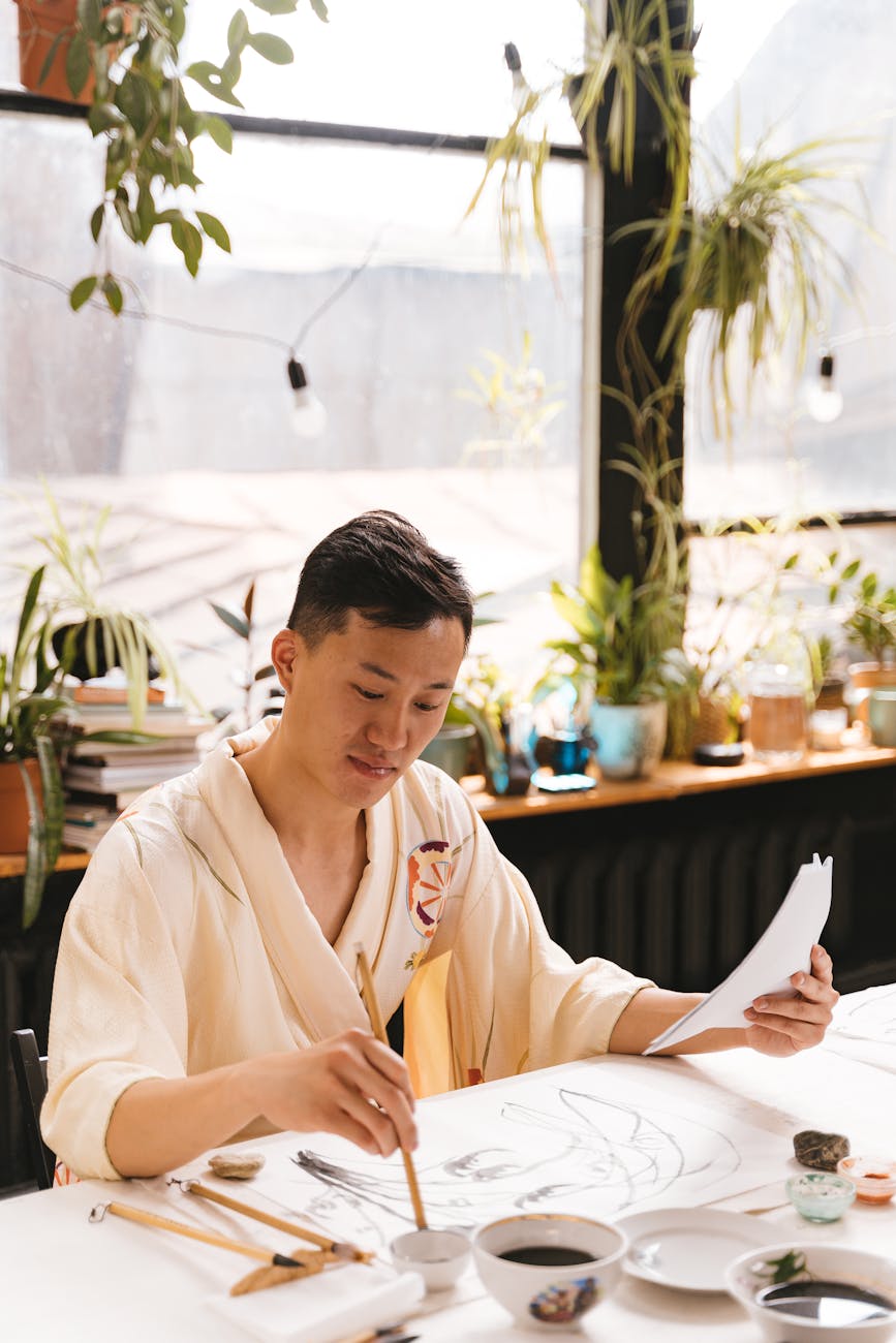 An adult man in a yukata painting creatively in a well-lit studio with art materials.