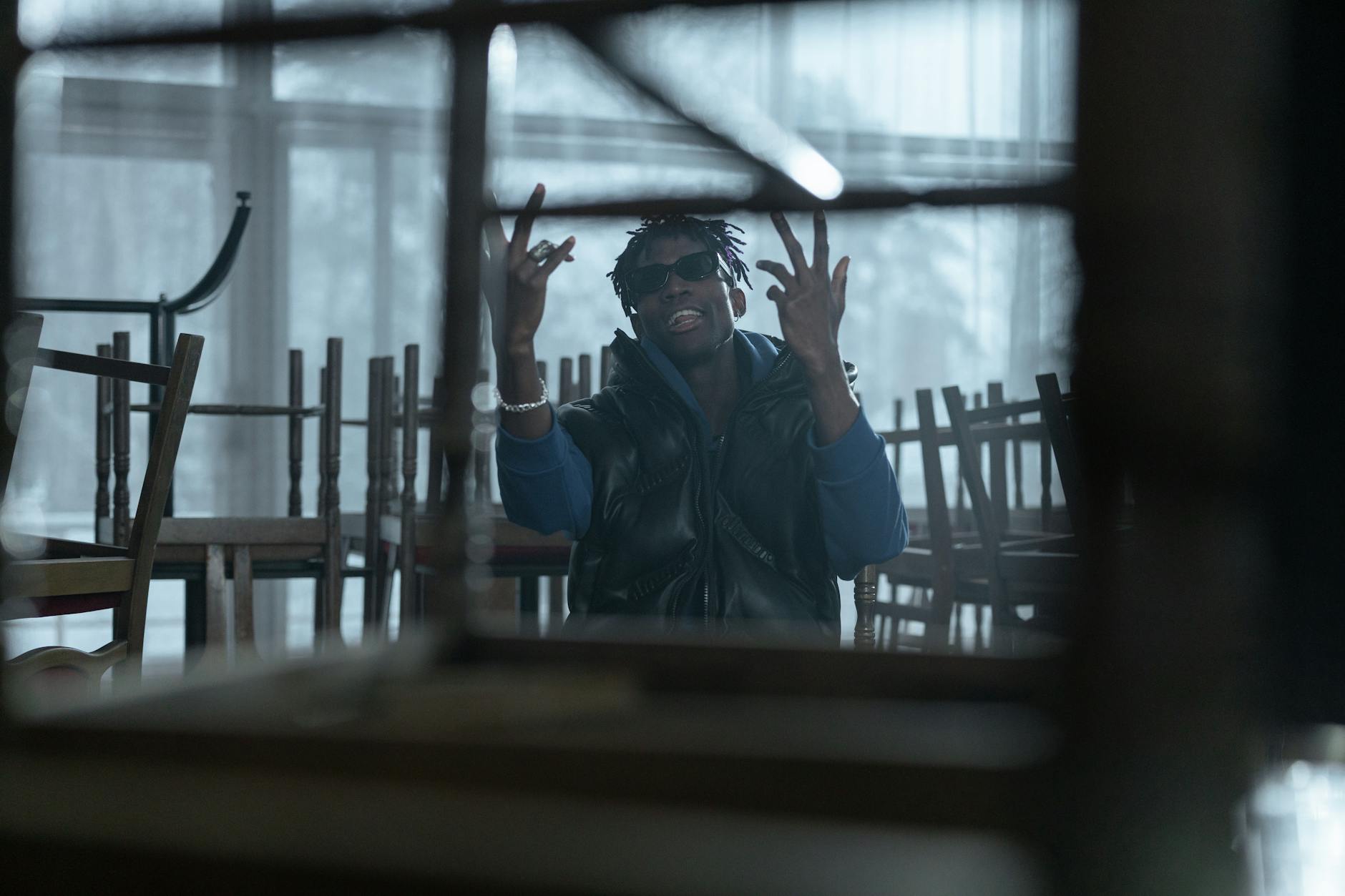 Young man in fashionable streetwear sitting stylishly among chairs inside a dimly lit room.