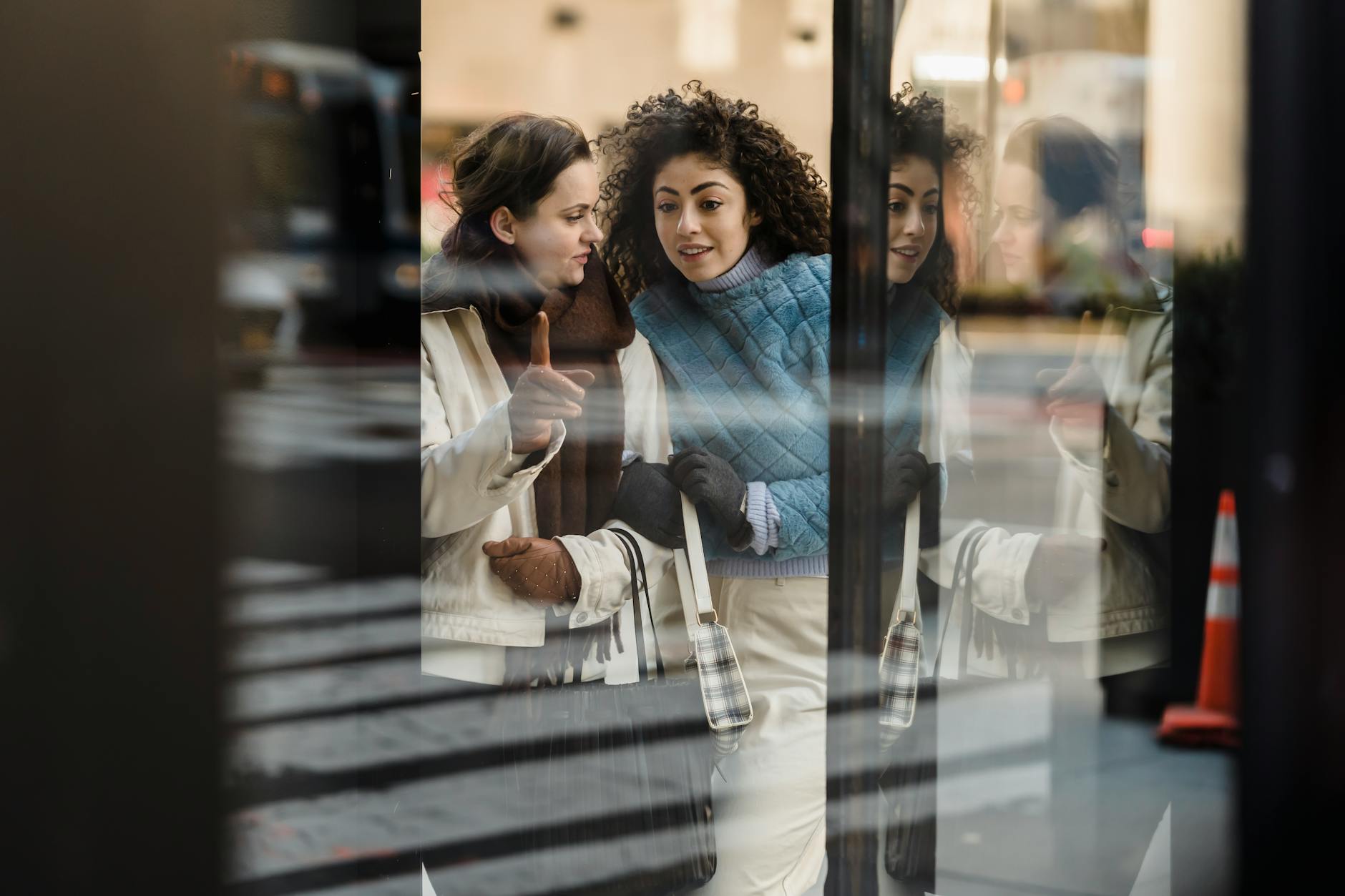 Through window of positive multiracial female friends standing near glass wall of modern shop while standing on street in city
