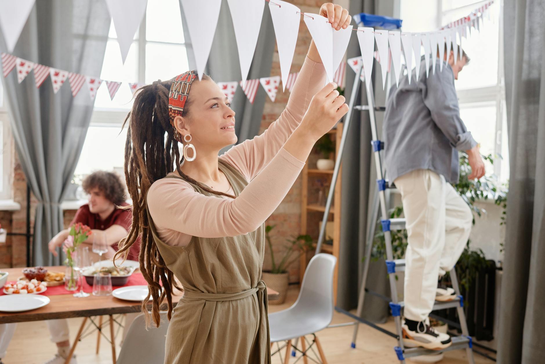 A woman with dreadlocks decorates for a festive indoor party with friends, setting up joyful bunting.