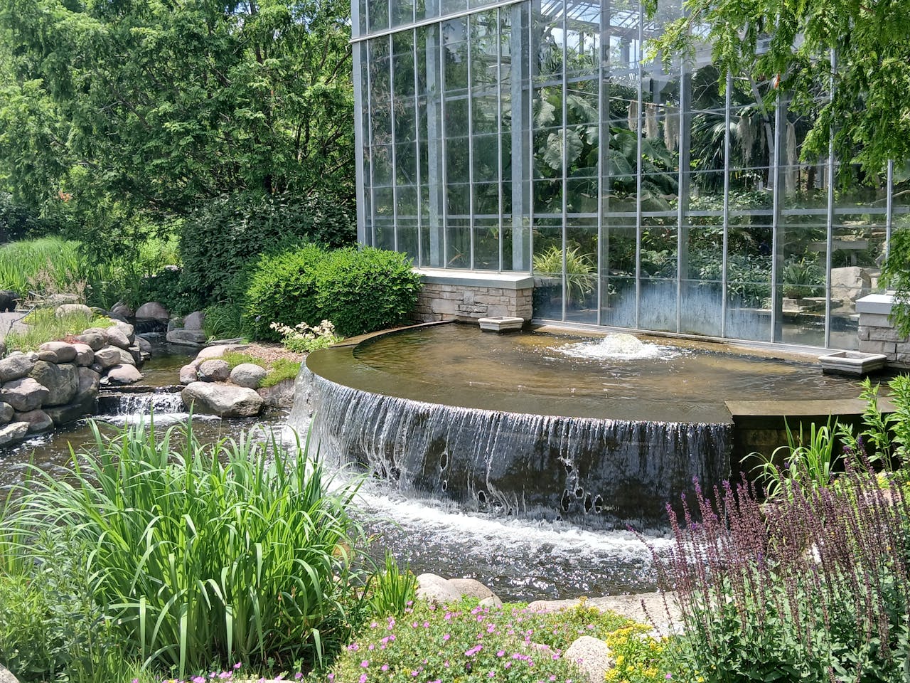 Beautiful outdoor waterfall beside a glass conservatory, lush with greenery in a peaceful setting.
