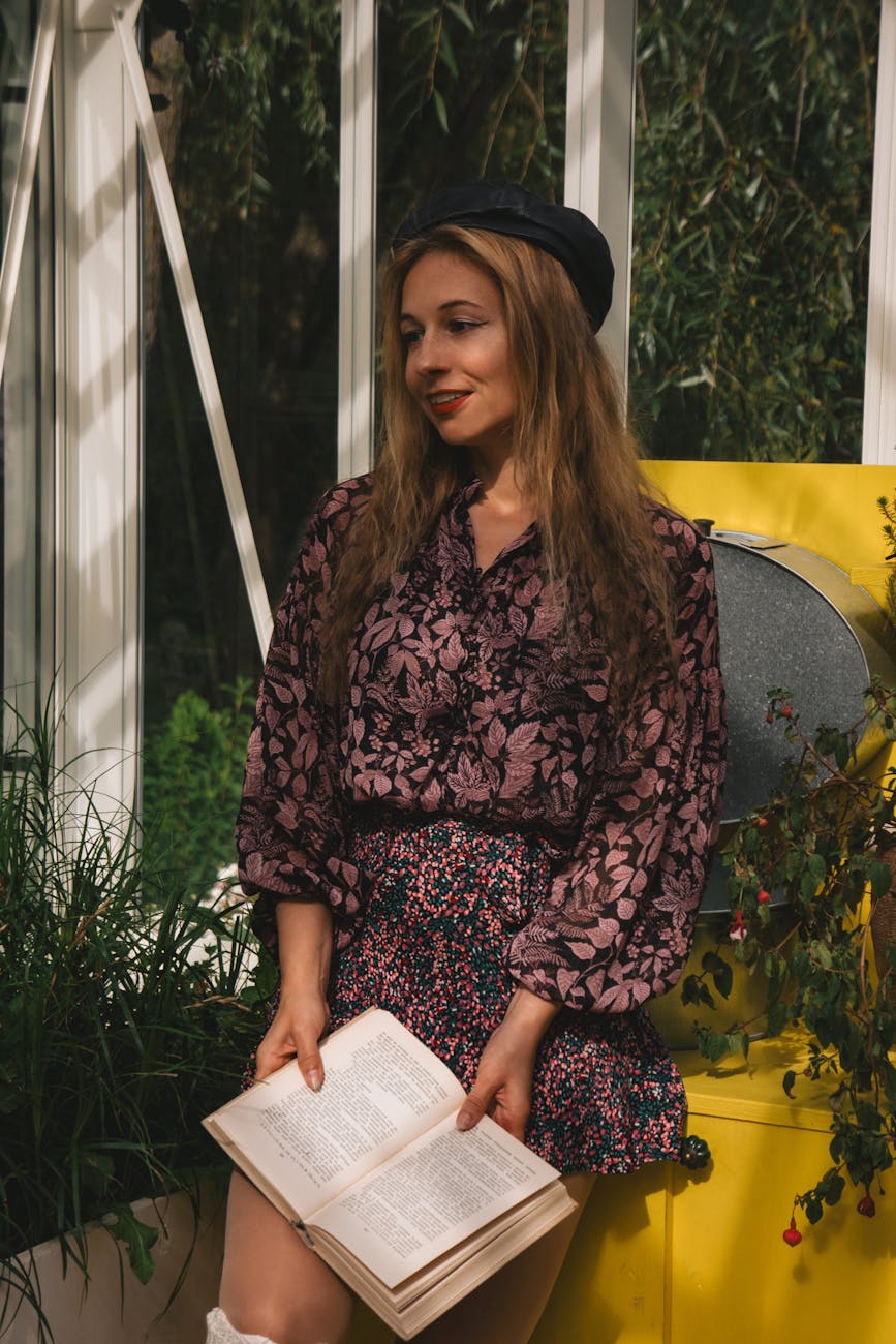 A stylish young woman enjoys reading in a sunlit conservatory filled with plants.