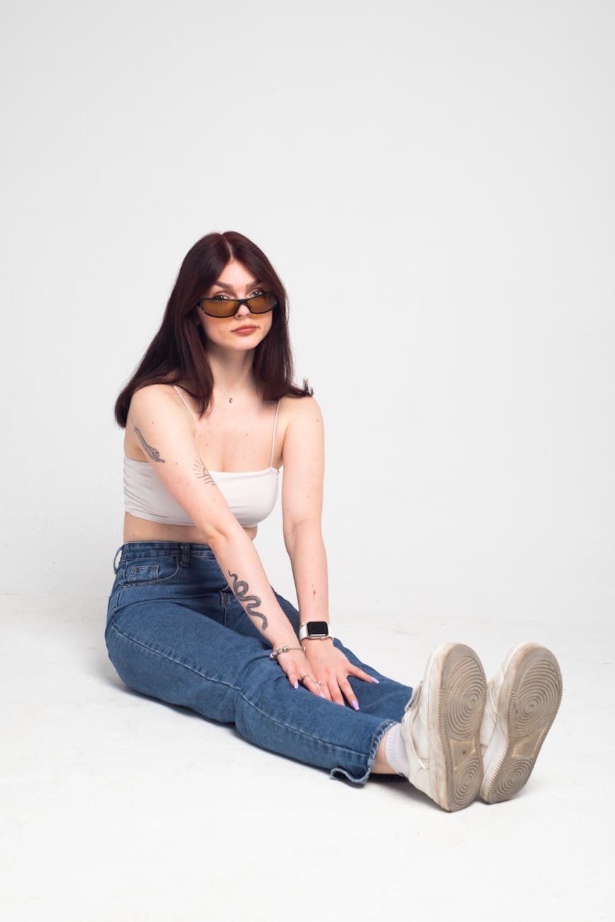 Trendy woman with tattoos sitting in casual jeans and sunglasses on a white background.